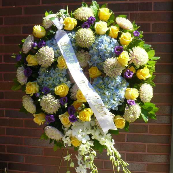 Standing funeral wreath with yellow roses, blue hydrangeas, and purple flowers