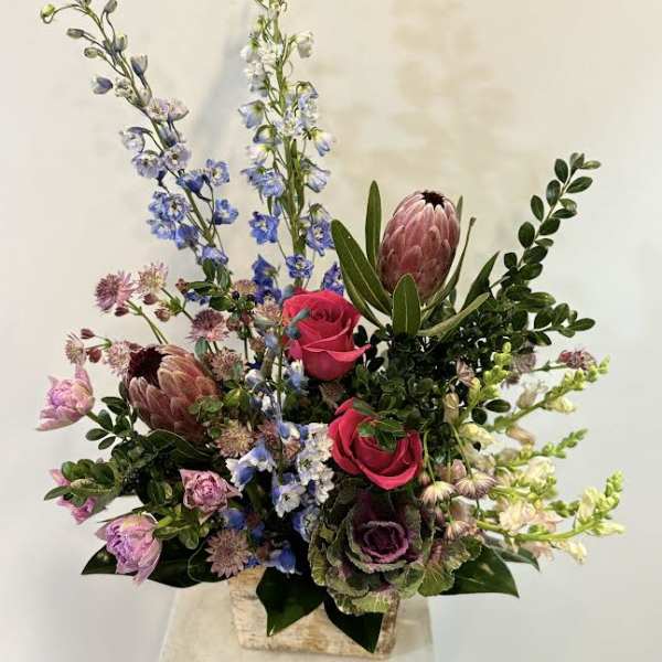 Tall mixed bouquet with pink roses, blue delphinium, and protea in a square vase