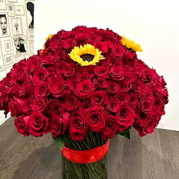 Large arrangement of red roses with yellow sunflowers in a clear glass cylinder vase with red ribbon.