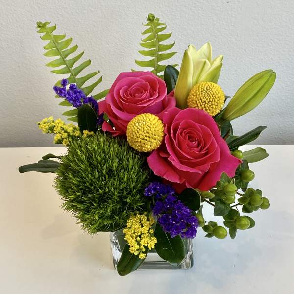 Pink roses in a clear square vase with yellow and purple accents