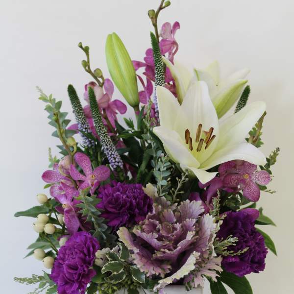 Tall arrangement of white lilies and purple flowers in a white ceramic cube vase with embossed heart.