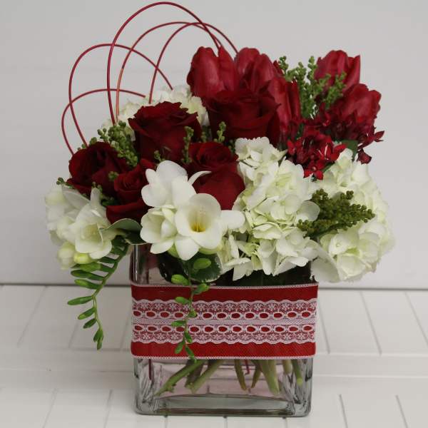 Low red and white flower arrangement with roses and hydrangeas in a square glass vase wrapped with red lace ribbon