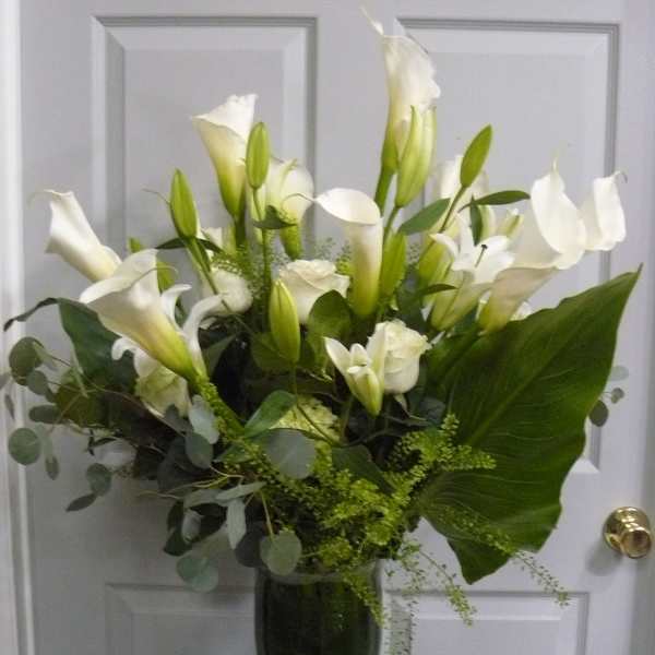 Tall arrangement of white calla lilies and roses with a large green leaf in a clear glass vase