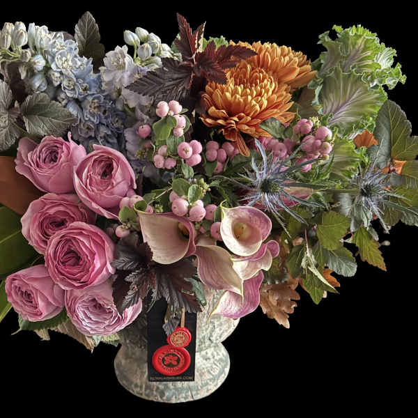 Lush arrangement with pink roses, mini calla lilies, orange mums, blue flowers and ornamental cabbage in a footed vase