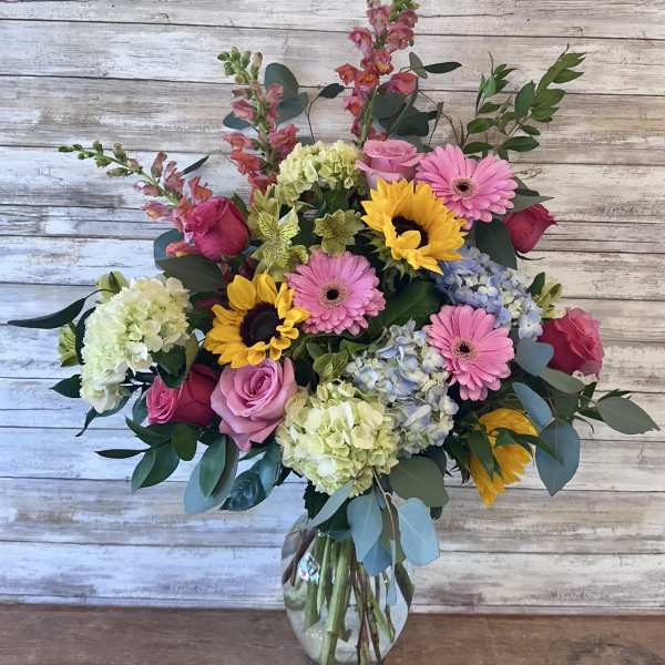 Mixed bouquet of pink roses, gerbera daisies, sunflowers, and hydrangeas in a glass vase