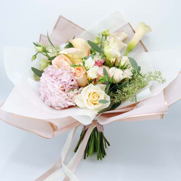 Hand-tied bouquet of cream roses, pink hydrangea, and pale calla lilies wrapped in blush paper.