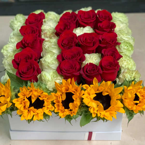 Red roses and white roses arranged in a white box with sunflowers along the front