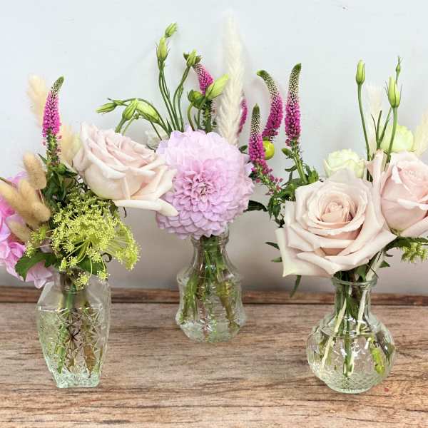 Trio of small glass bud vases with blush roses, a pink dahlia, and delicate accent flowers.