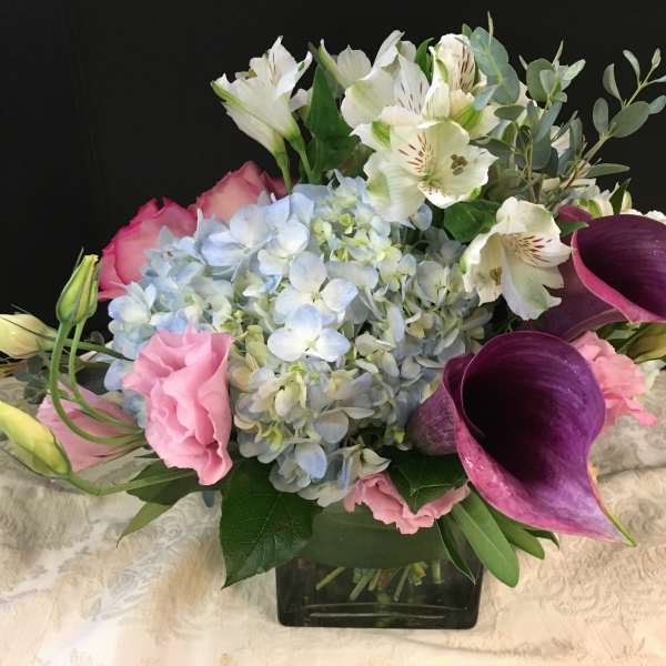 Low arrangement with blue hydrangea, purple calla lilies and pink blooms in a glass cube vase