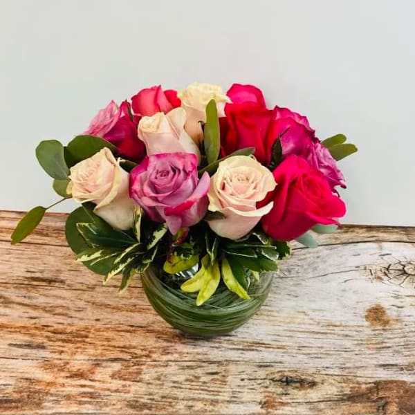 Round arrangement of pink, cream, and lavender roses in a clear glass bowl vase