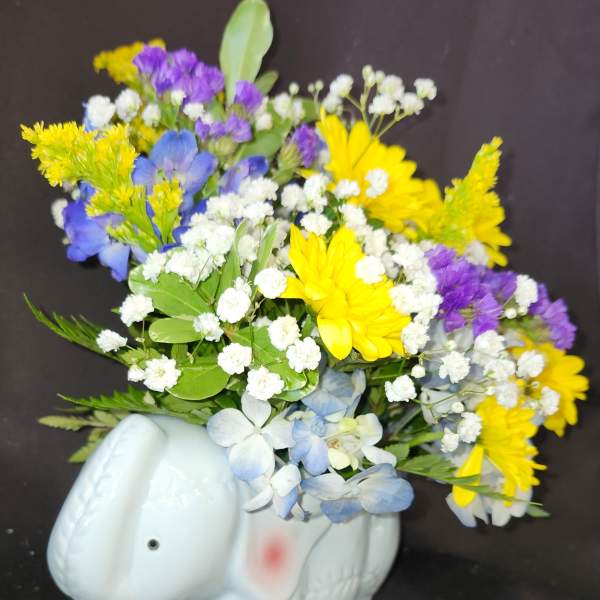 Mixed yellow, purple, white, and blue flowers in a white elephant planter