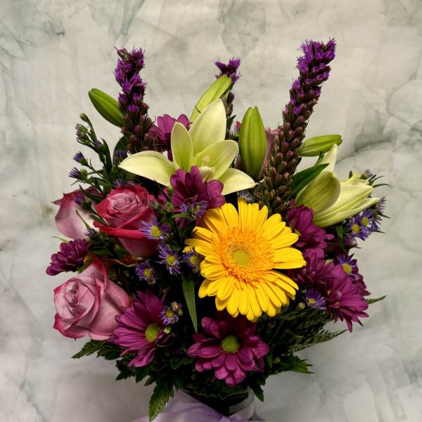 Mixed bouquet of roses, lilies, and daisies in a black vase with a lavender ribbon