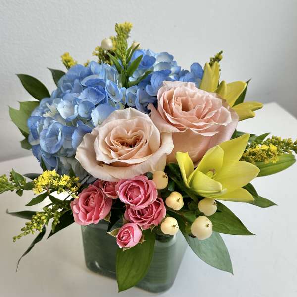 Bouquet of blue hydrangeas, peach roses, pink spray roses, and yellow lilies in a glass vase