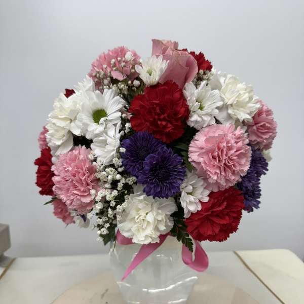 Mixed pink, white, red, and purple flowers in a clear glass vase with a pink ribbon