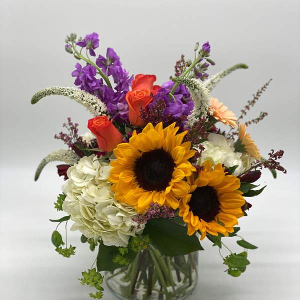 Bouquet of sunflowers, roses, hydrangea, and purple blooms in a glass vase