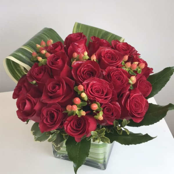 Red roses arranged in a clear square vase with green leaf wrap