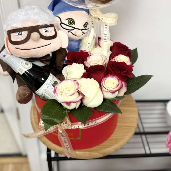 Gift set with red and white roses in a red box, two plush dolls, a bottle, and a clear anniversary balloon.