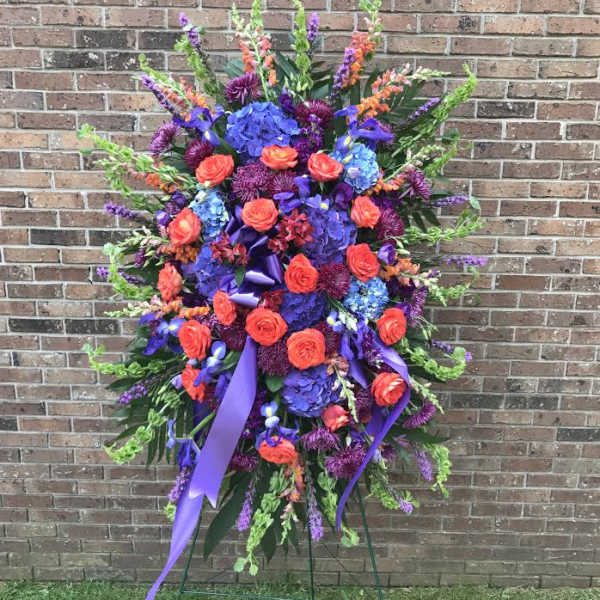 Tall standing spray of purple, blue, and orange flowers with ribbons on a metal easel