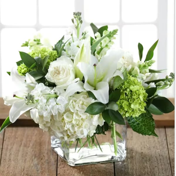 White lilies and roses arranged in a clear square vase with green accents