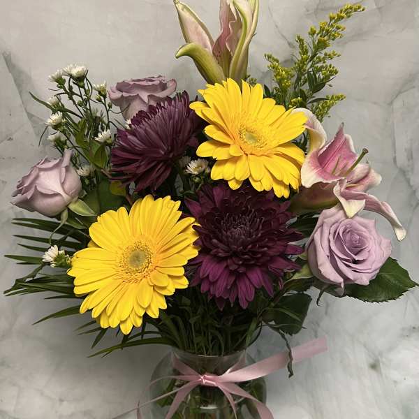 Bouquet of yellow gerbera daisies, purple chrysanthemums, and lavender roses in a glass vase