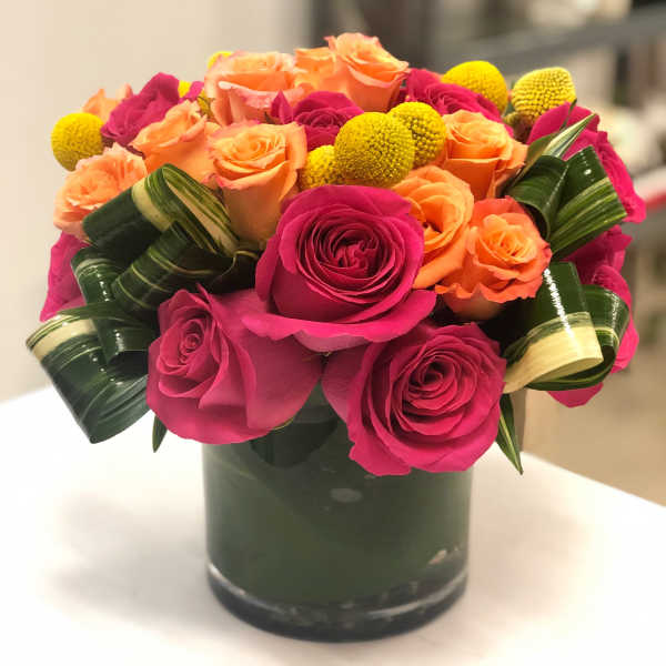 Compact arrangement of hot pink and orange roses with yellow accents in a glass cylinder vase
