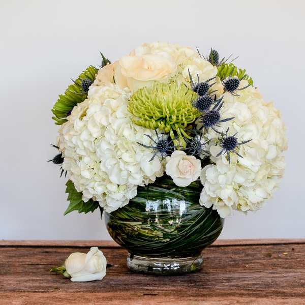 Low round arrangement of white hydrangeas, roses, and green mums in a leaf-wrapped glass bowl vase
