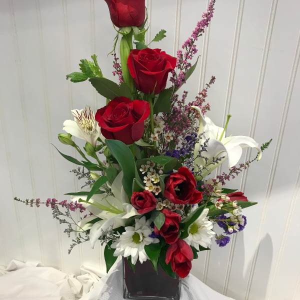 Tall arrangement of red roses, white lilies, and daisies in a square vase