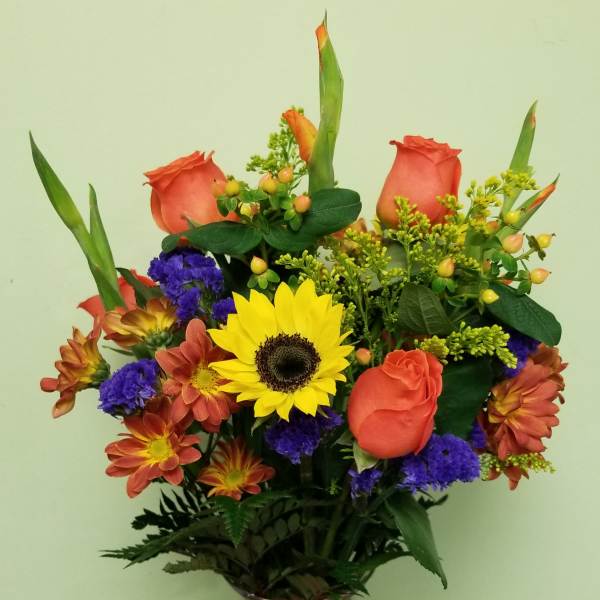 Mixed bouquet of orange roses, sunflowers, and purple flowers in a glass vase