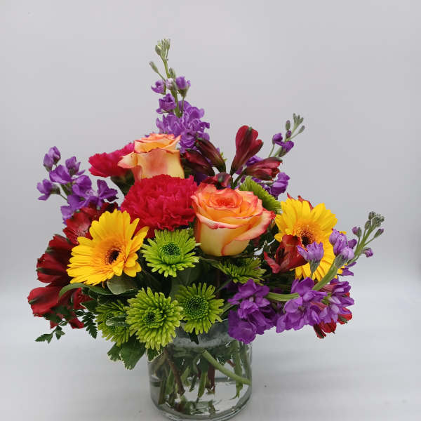 Mixed bouquet of roses, gerbera daisies, carnations, and purple blooms in a glass vase