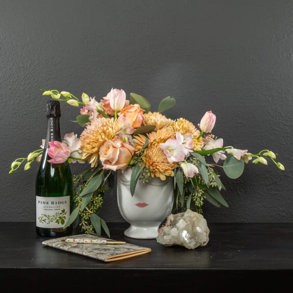 Floral arrangement in a white vase with a sparkling wine bottle and notebook