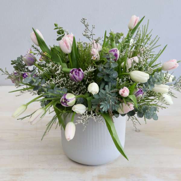 Low white ceramic pot with pink, purple, and white tulips arranged with airy greenery.