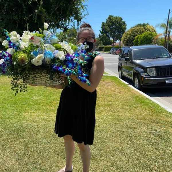 Person in a black dress holding a large basket of white, blue, and purple flowers on a lawn