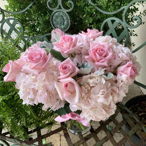 Low glass vase of light pink roses and hydrangeas arranged in a compact mound on a metal bench
