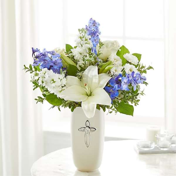 White lilies and blue flowers arranged in a cream vase with a cross emblem