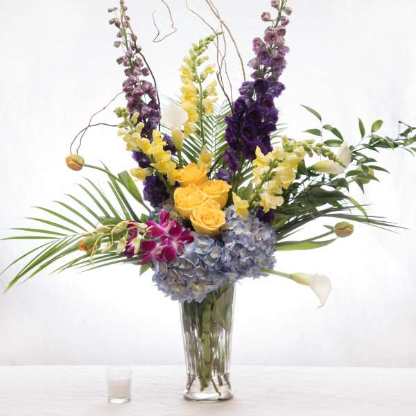 Tall mixed arrangement with blue hydrangeas, yellow roses, snapdragons, orchids, and calla lilies in a glass vase.