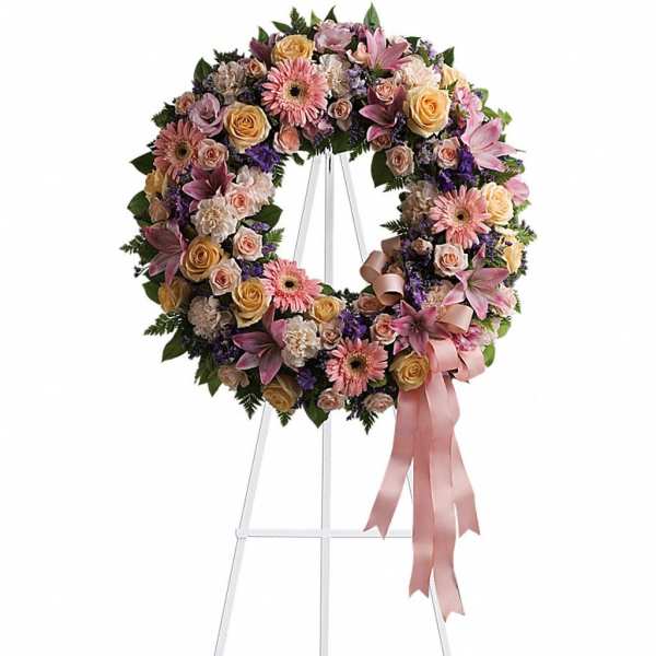 Pastel wreath of roses, lilies, gerbera daisies, and carnations on a white easel with trailing pink ribbon.