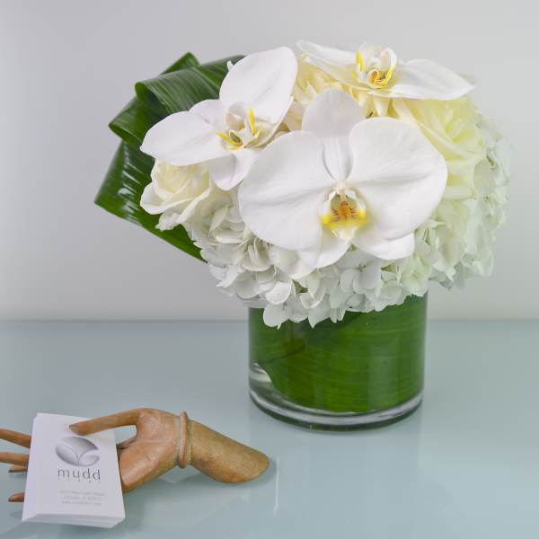 White orchids and hydrangeas in a glass vase with broad green leaves