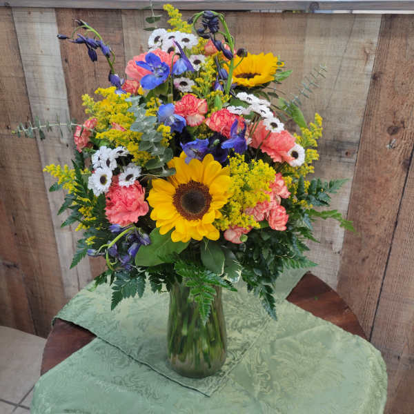 Mixed bouquet with sunflowers, daisies, carnations, and blue flowers in a glass vase