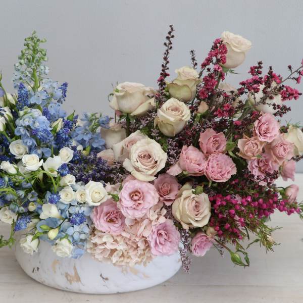 Low centerpiece with blue and white blooms on one side and pink and cream roses on the other in a white bowl