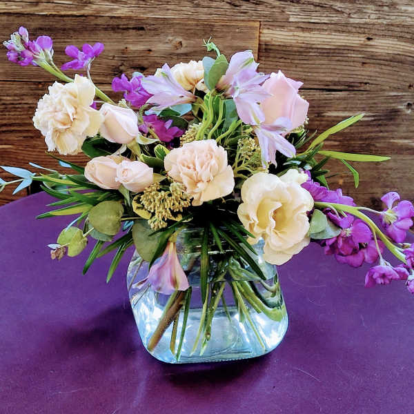 Low arrangement of peach roses and carnations with purple flowers in a clear glass cube vase