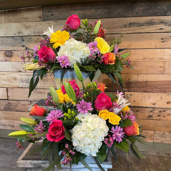 Two colorful floral arrangements in metal containers with roses, hydrangeas, lilies, and daisies