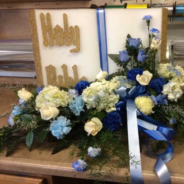 Religious floral tribute with white and blue flowers arranged before a Holy Bible plaque.