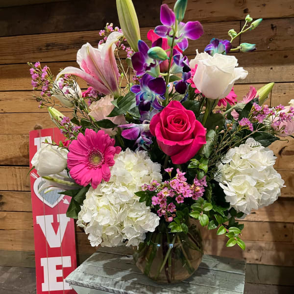 Mixed arrangement of pink, white, and purple flowers in a glass bowl vase on a rustic wooden bench