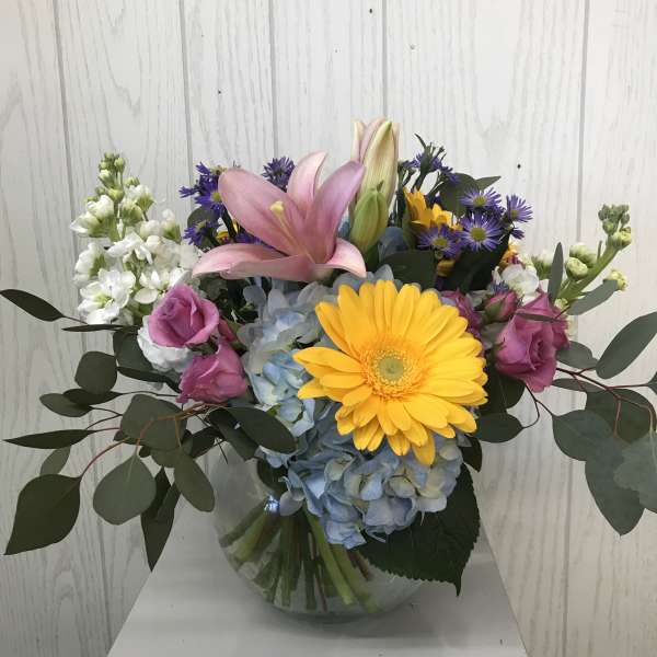 Mixed bouquet with pink lilies, yellow gerbera daisy, blue hydrangea, and purple blooms in a glass vase
