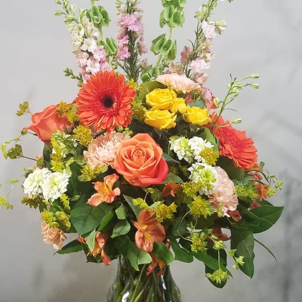 Tall mixed bouquet with orange gerbera daisies, roses, yellow roses, and green bells of Ireland in a clear glass vase