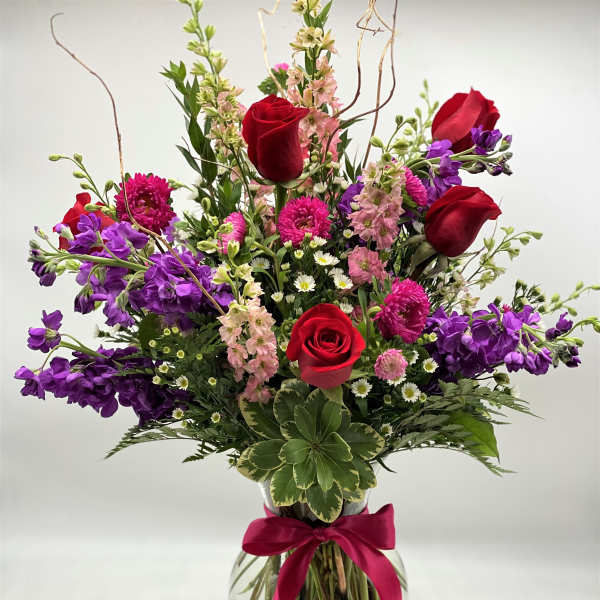 Mixed bouquet of red roses, purple blooms, and pink flowers in a glass vase with ribbon