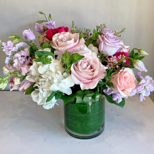 Pink and lavender roses arranged with white hydrangeas in a green glass vase.