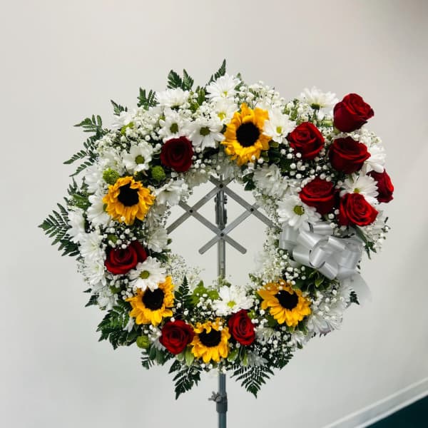 Circular floral wreath with red roses, white daisies, and sunflowers on a stand
