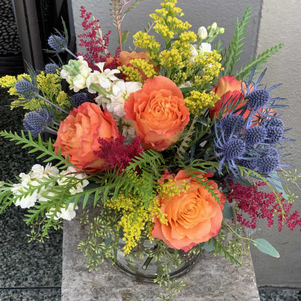Bouquet of orange roses, white blooms, and blue thistles in a glass vase