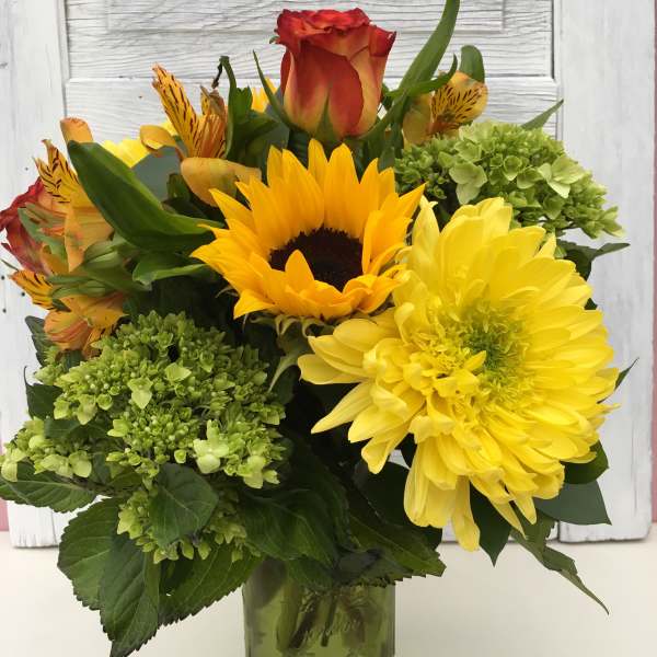 Bouquet of sunflowers, yellow daisies, roses, and green hydrangeas in a glass vase.